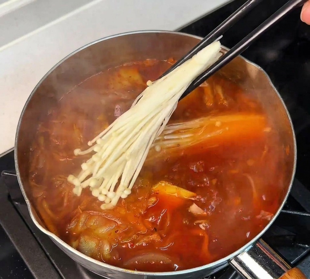 끓는 김치찌개 속에 팽이버섯 묶음을 젓가락으로 넣고 있는 모습
