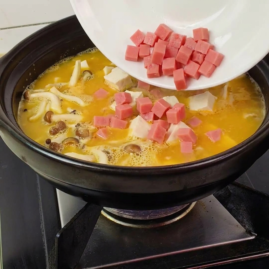 노란 국물이 담긴 뚝배기에 두부, 햄, 버섯, 완두콩을 넣는 모습