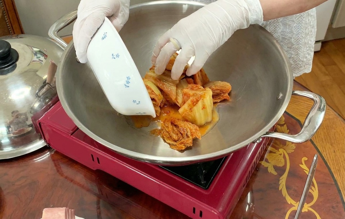 빨간 휴대용 버너 위에 놓인 냄비 중앙에 썰어둔 신김치를 담는 모습.