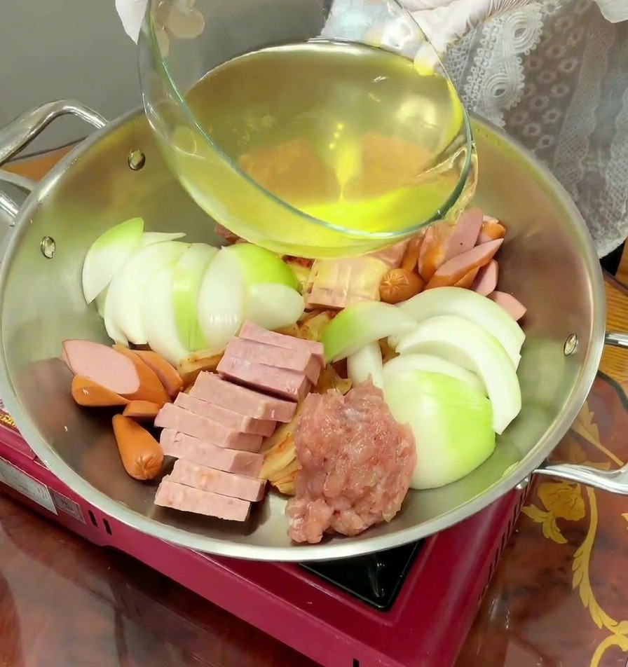 정갈하게 담긴 부대찌개 재료 위로 맑고 노란 육수를 붓는 모습.