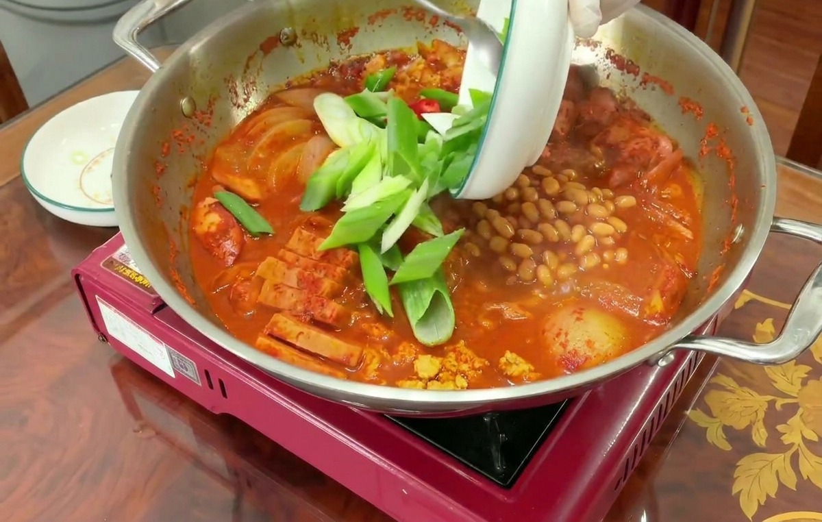 끓고 있는 부대찌개 위로 신선한 대파와 팽이버섯을 풍성하게 올리는 모습.