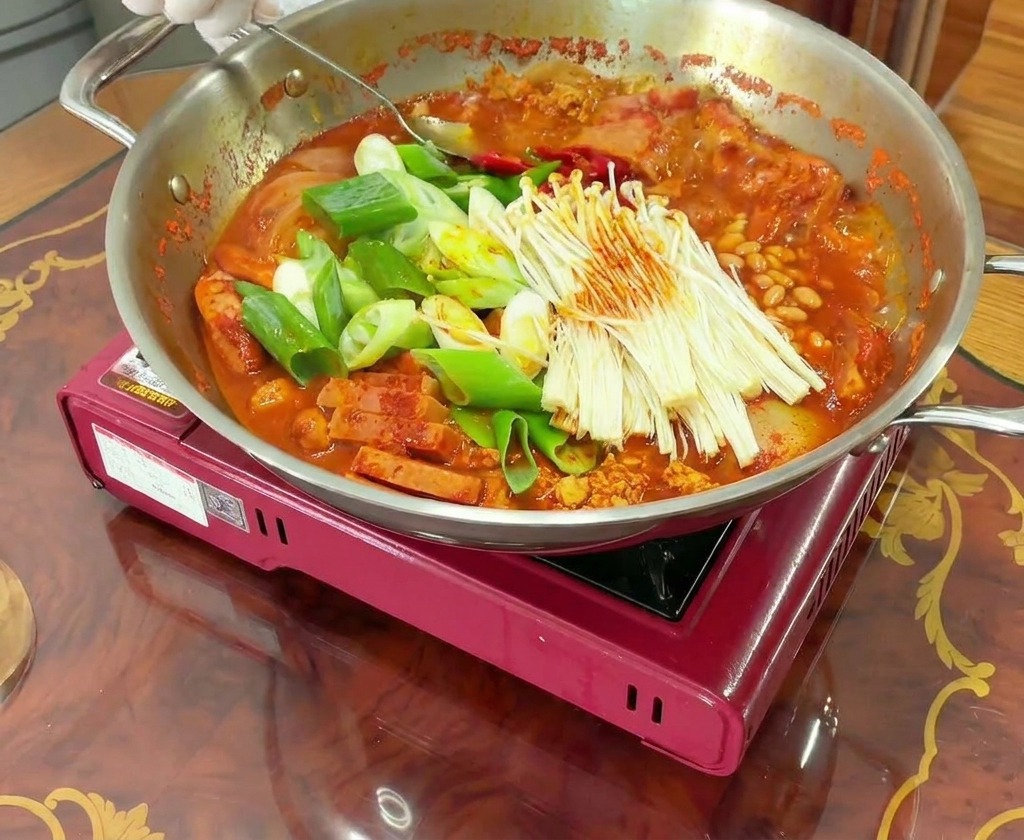 국자로 끓는 부대찌개의 고기와 채소, 빨간 국물을 고루 섞어주는 모습.