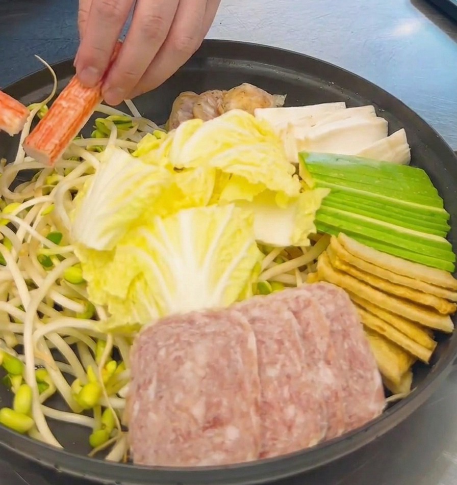 배추, 스팸, 두부가 담긴 냄비에 선명한 주황색 게맛살을 채워 넣는 손.