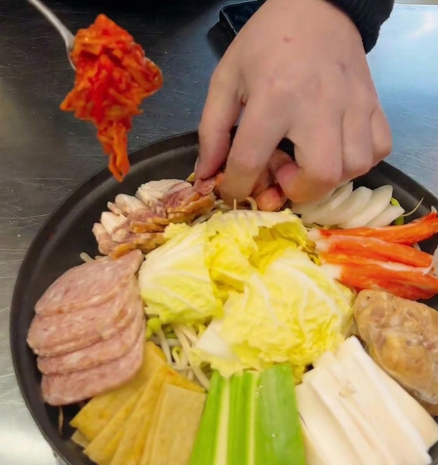 소시지와 채소들이 담긴 냄비 중앙에 수저로 잘 익은 김치를 듬뿍 올리는 모습.