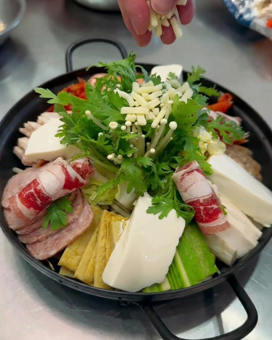 완성된 부대찌개 냄비 중앙의 채소 위에 하얀 치즈 가루를 뿌리는 근접 촬영.