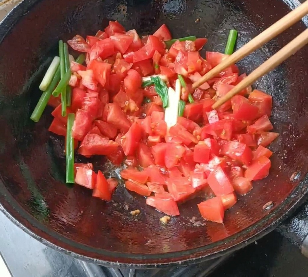 검은색 무쇠 웍에서 나무 젓가락으로 다진 토마토와 대파를 볶는 모습.