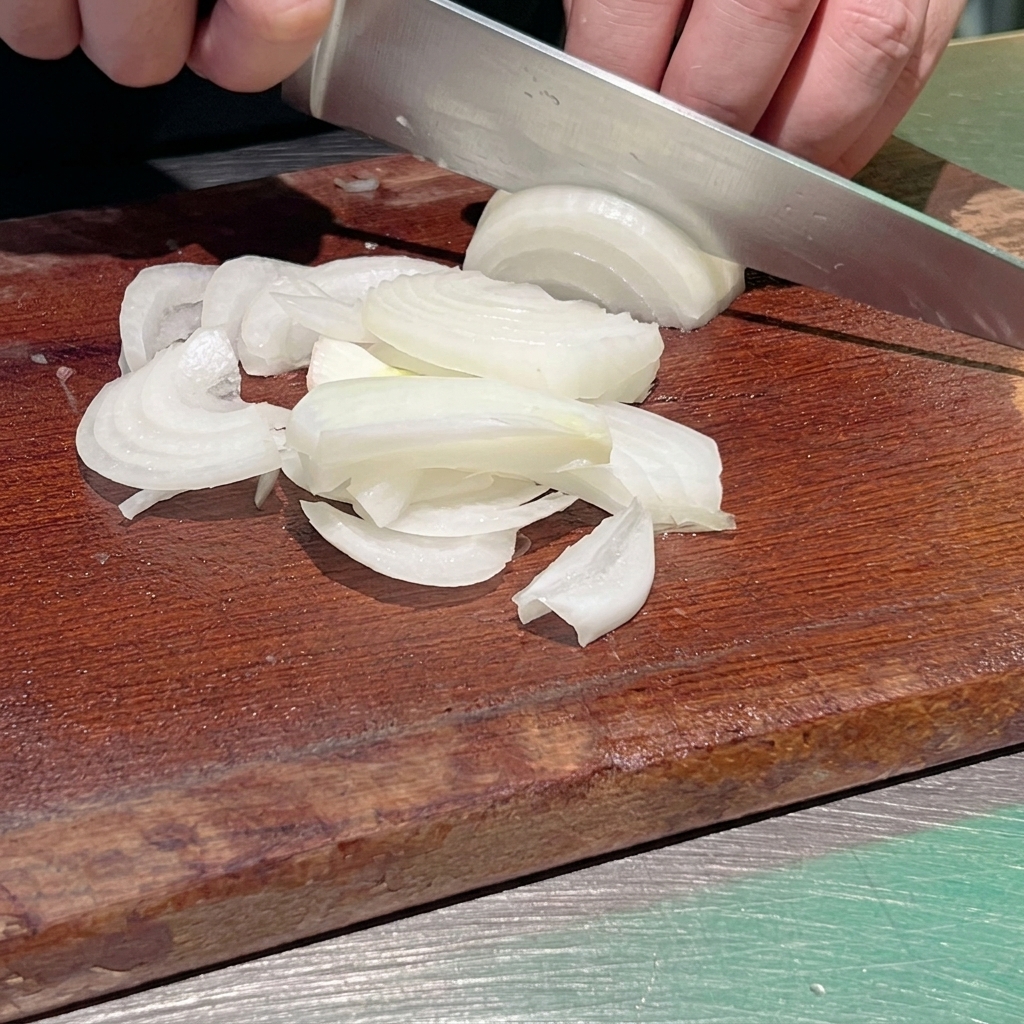 어두운 나무 도마 위에서 셰프가 날카로운 주방 칼로 흰 양파를 얇게 채썰고 있는 근접 사진.