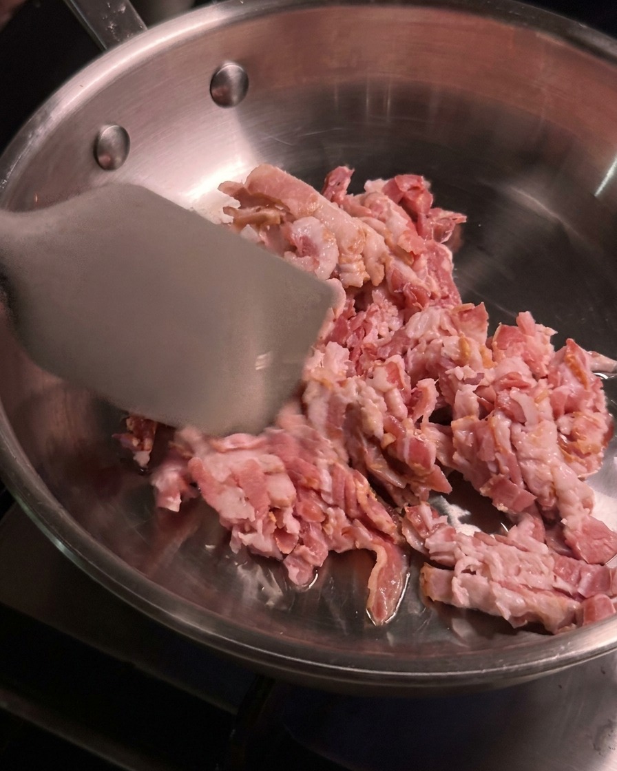 달궈진 스테인리스 팬 안에서 금속 뒤집개로 잘게 썬 생베이컨을 볶으며 익히는 모습.