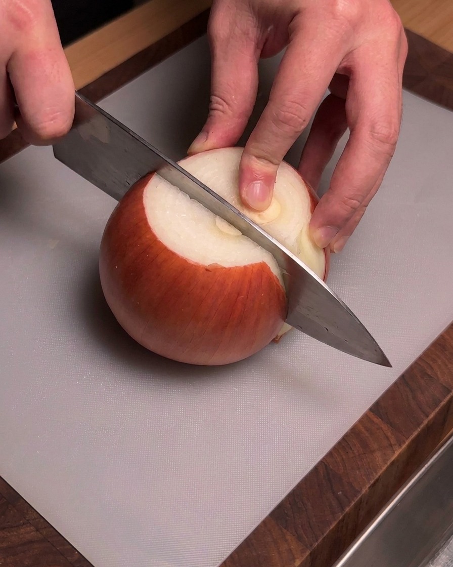 회색 도마 위에서 껍질을 벗긴 황양파를 셰프 나이프로 조심스럽게 써는 양손의 모습.
