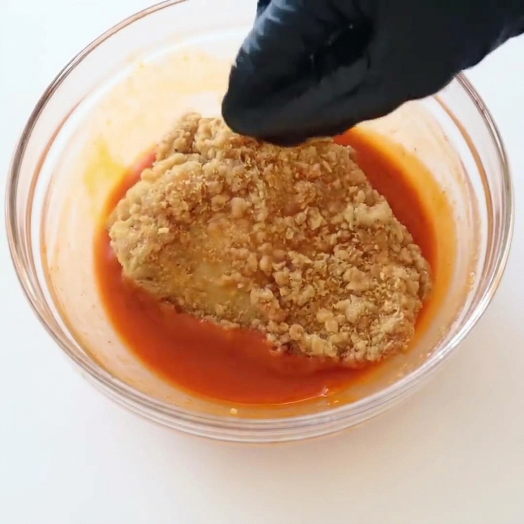 검은색 장갑을 낀 손이 밝은 붉은색 버팔로 소스가 담긴 유리 볼에 노릇하고 바삭하게 튀겨진 치킨 필레를 담그는 모습.