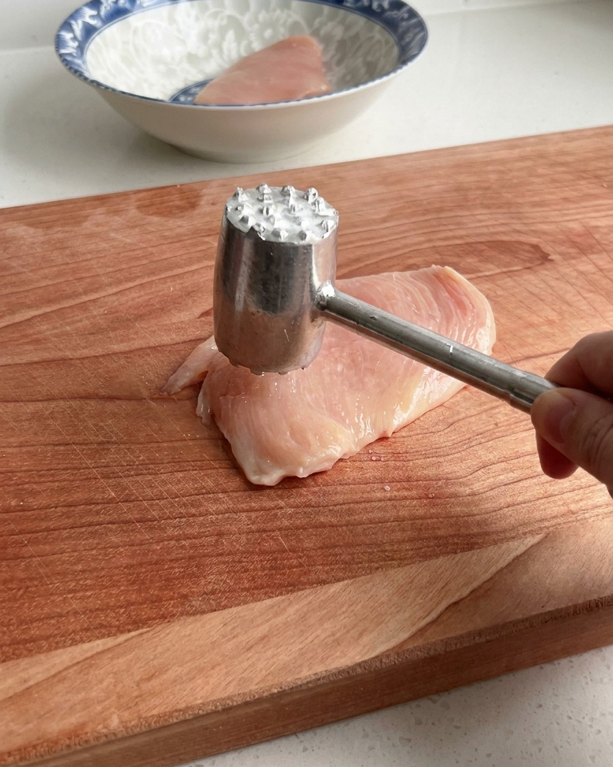 나무 도마 위에서 고기 망치로 닭가슴살을 부드럽게 두드리고 있는 모습.