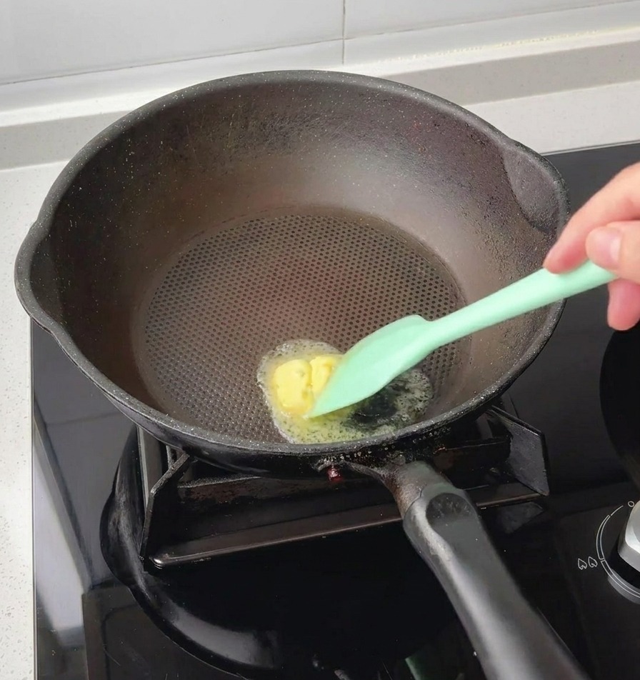 달궈진 검은색 팬 중앙에서 실리콘 주겁으로 네모난 노란 버터를 녹이는 모습.