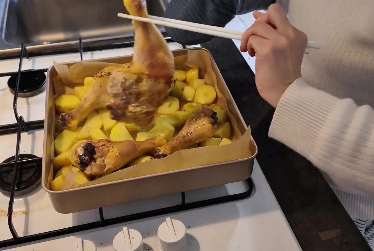 오븐 장갑을 낀 사람이 젓가락을 이용해 감자가 가득 담긴 뜨거운 오븐 팬 속의 닭다리를 조심스럽게 뒤집는 모습