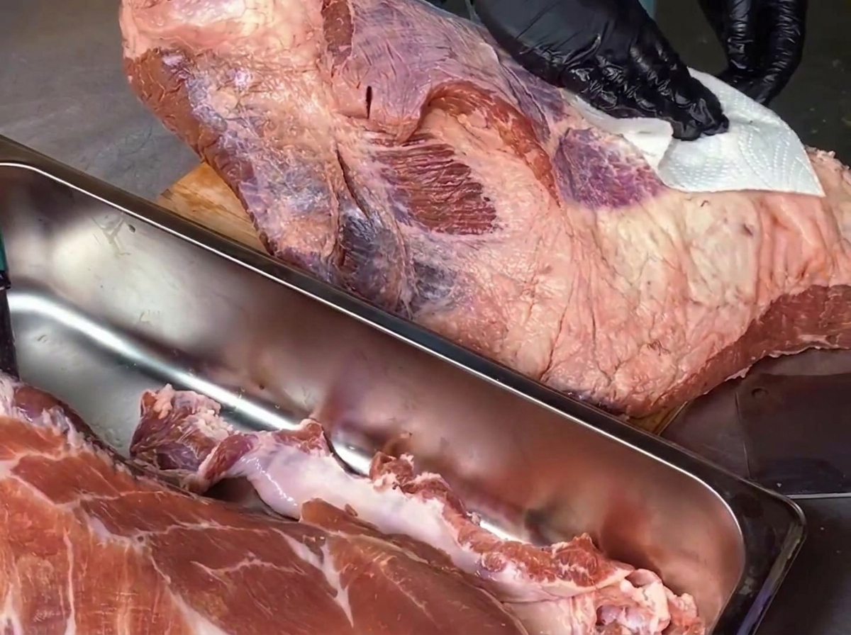 검은색 위생 장갑을 낀 셰프가 나무 도마 위에 놓인 거대한 생 차돌양지 고기의 표면을 흰색 키친타월로 닦아내고 있는 모습. 앞쪽에는 고기 자투리가 담긴 스테인리스 쟁반이 보인다.