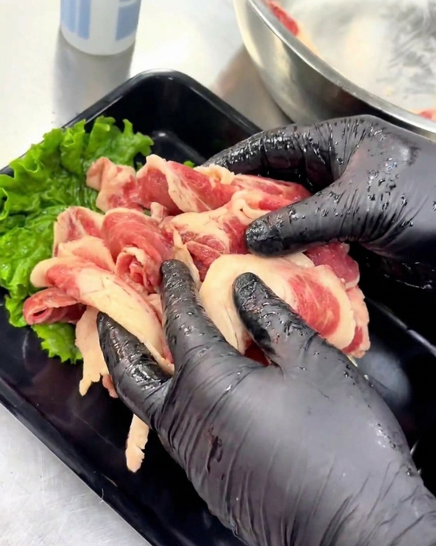 초록색 상추 잎으로 장식된 검은색 서빙 트레이에 양념된 생고기 슬라이스를 가지런히 올려놓는 장갑 낀 손.