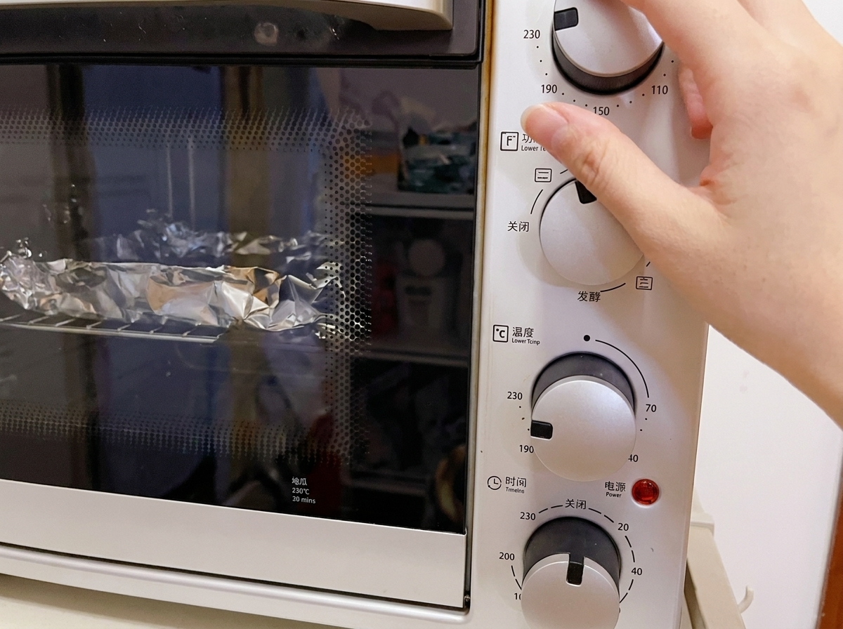 하얀색 미니 오븐의 온도 조절 다이얼을 230°C로 맞추는 손과 내부 그릴 위 호일 뭉치들