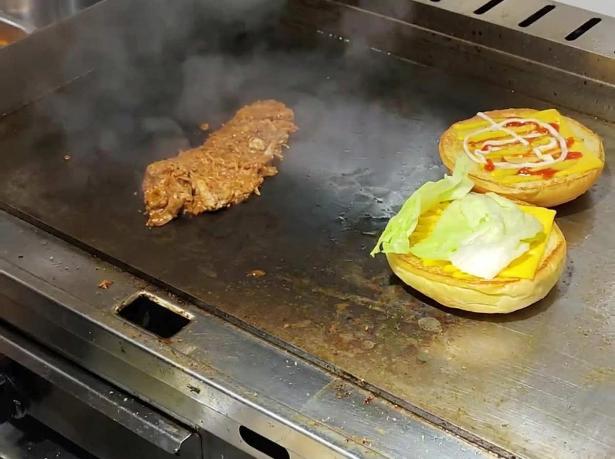 김이 나는 철판 위에서 노릇하게 구워진 버거 번에 치즈와 양상추를 올리고 그 옆에 따뜻한 풀드포크가 함께 놓여 있는 모습