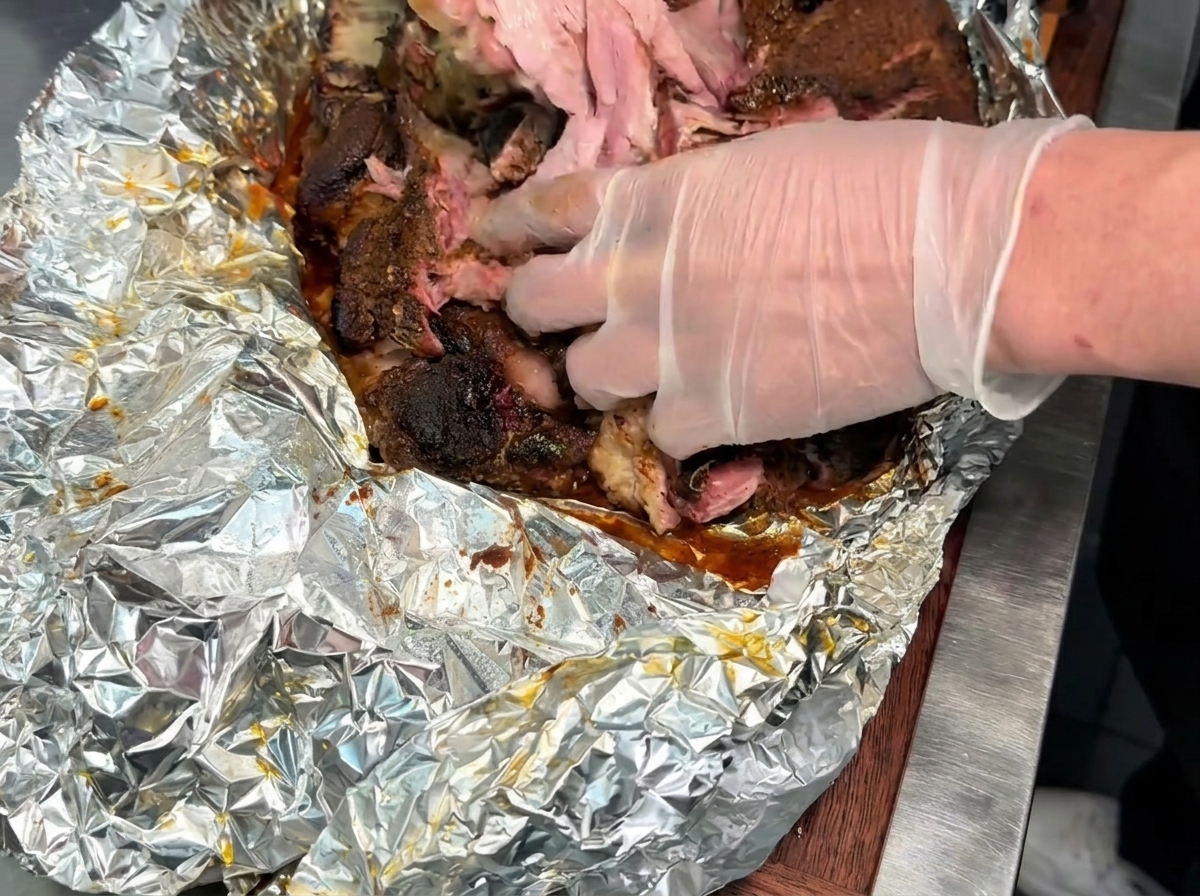장갑을 낀 손으로 호일이 깔린 바닥 위에서 잘 익은 부드러운 훈제 돼지고기 덩어리를 결대로 잘게 찢는 모습.