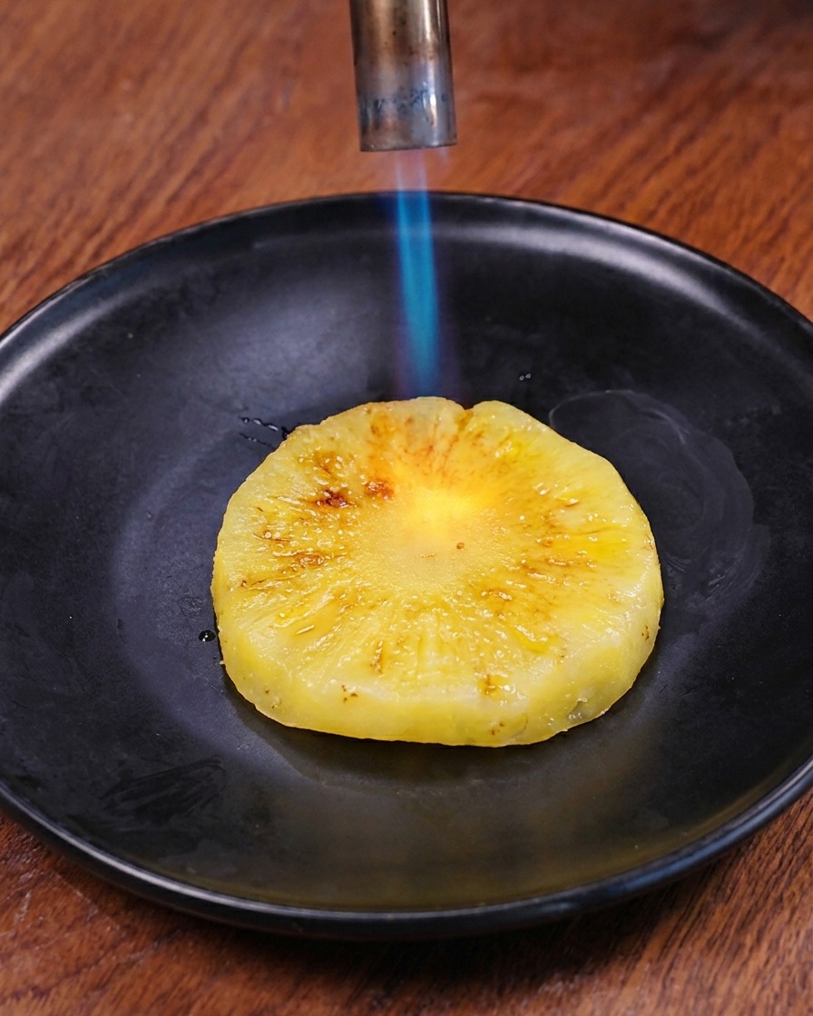 어두운 접시 위에 놓인 신선한 링 모양 파인애플 표면을 주방용 토치의 푸른 불꽃으로 그을리는 모습.