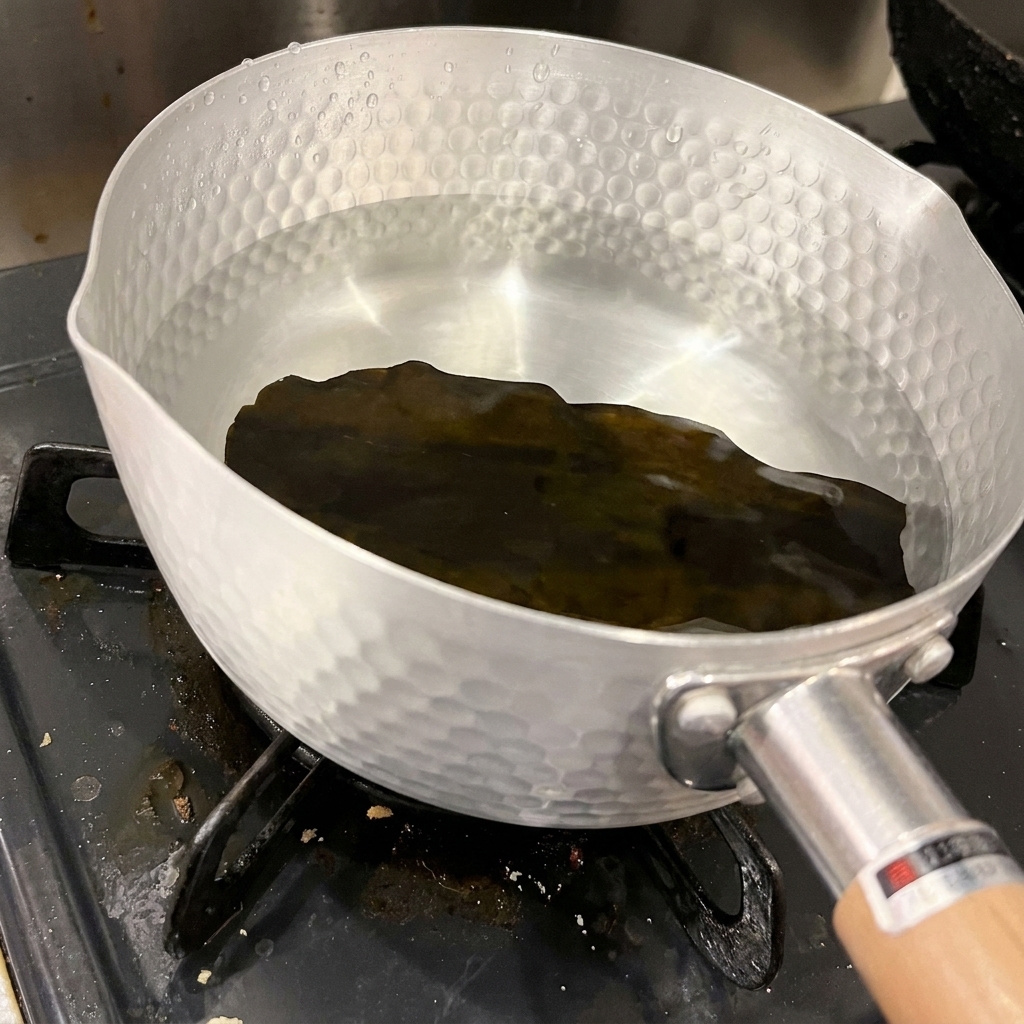 망치로 두드린 자국이 있는 알루미늄 냄비 바닥에 맑고 찬물과 함께 담겨 있는 진한 색의 건다시마 한 조각.