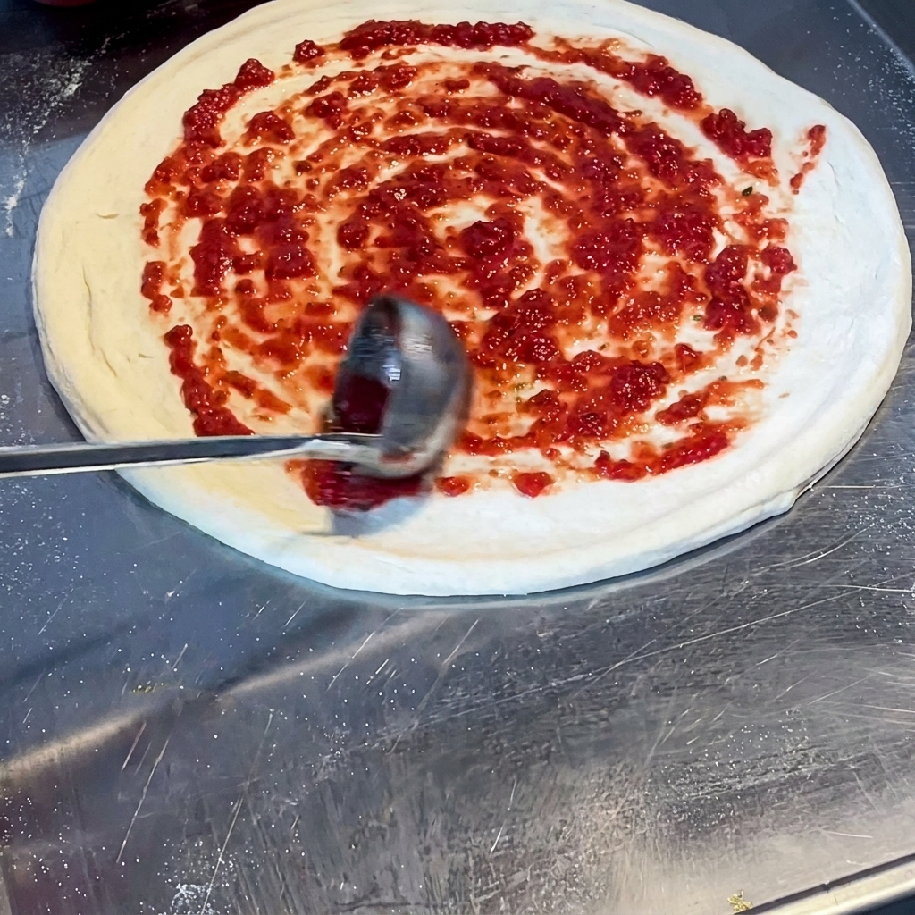 국자를 사용하여 도우 위에 토마토 소스를 나선형으로 바르는 모습.