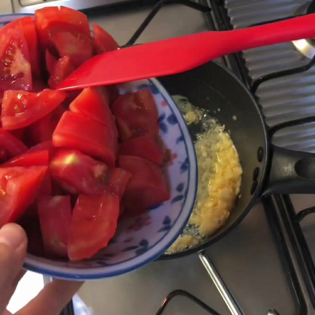 버터와 다진 마늘이 녹아 있는 검은색 냄비에 토마토 조각을 넣는 모습.