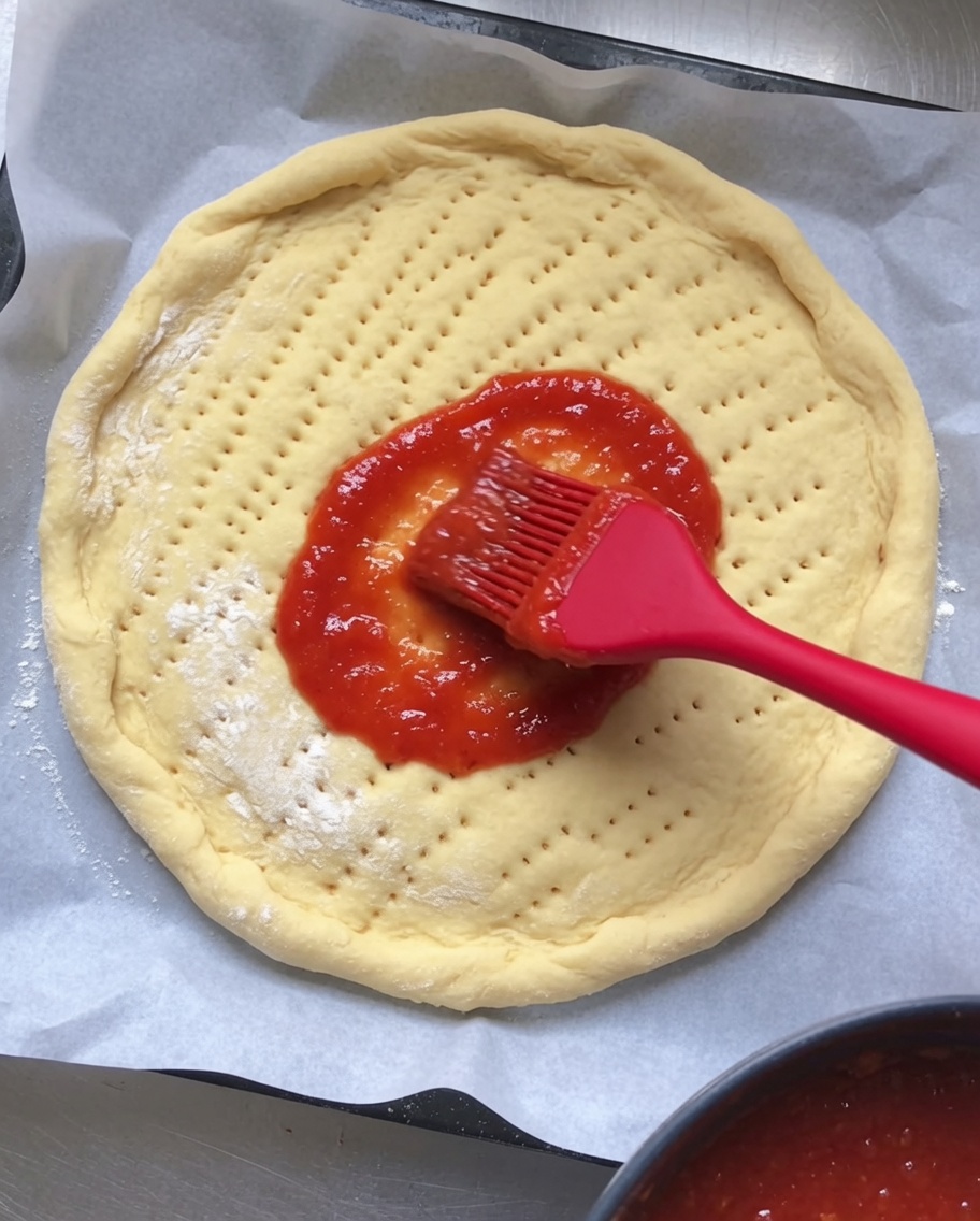 구멍이 난 둥근 생피자 도우 위에 붉은 토마토 소스를 고르게 펴 바르는 모습.