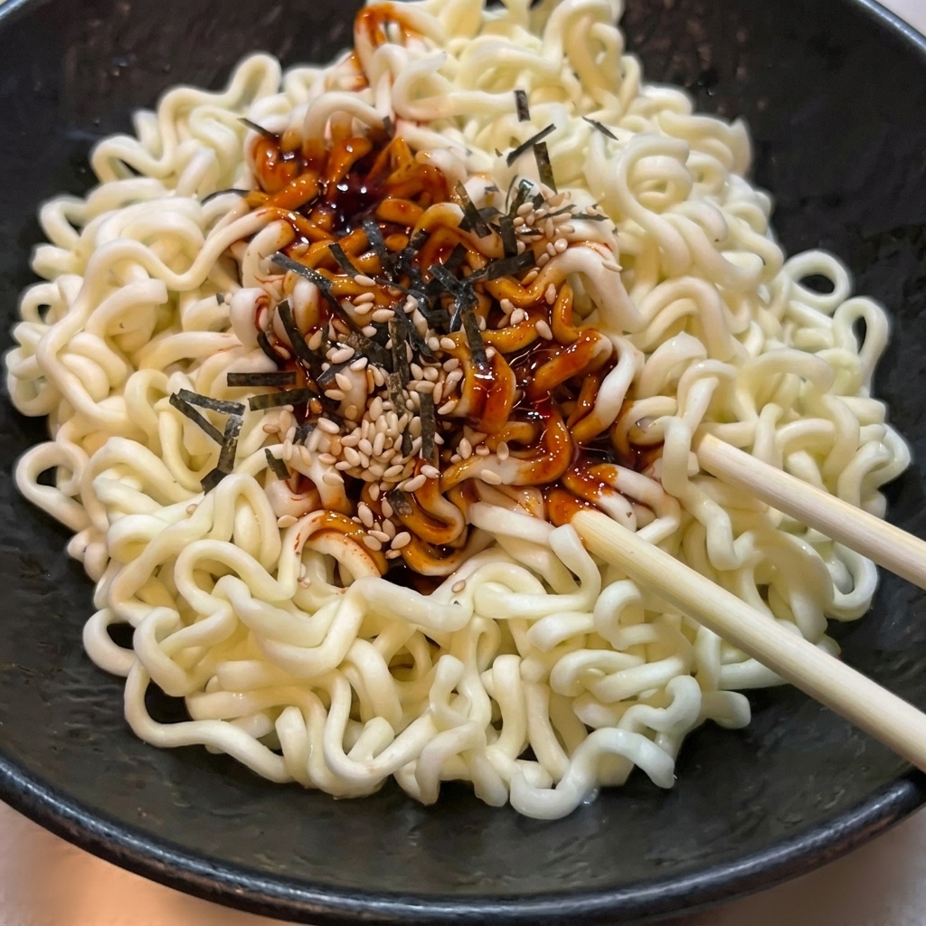 질감 있는 검은 그릇에 담긴 웨이브 라면 면발을 나무 젓가락으로 매운 소스, 깨, 김가루와 함께 버무리는 모습.