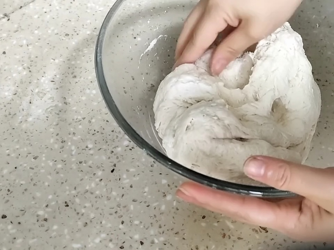 유리 볼 안에서 거친 반죽 덩어리를 손으로 치대며 반죽하는 모습.
