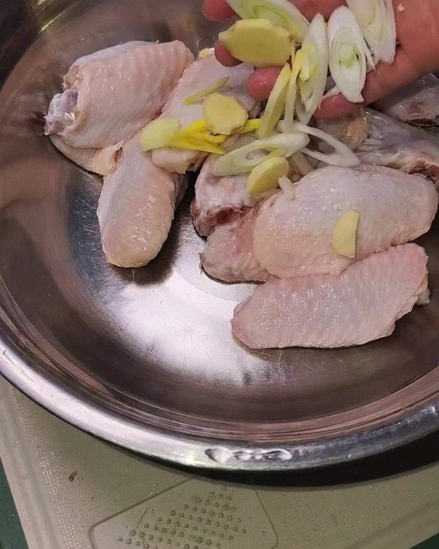 스테인리스 볼에 담긴 손질된 닭날개 위에 슬라이스한 생강, 마늘, 대파가 올려져 있는 모습.