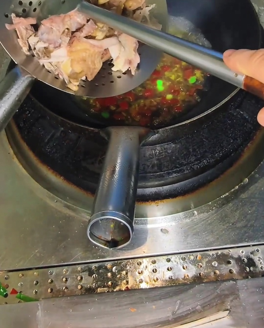 매운 육수가 끓고 있는 웍에 금속 구멍 국자로 찢은 닭고기를 넣는 모습.
