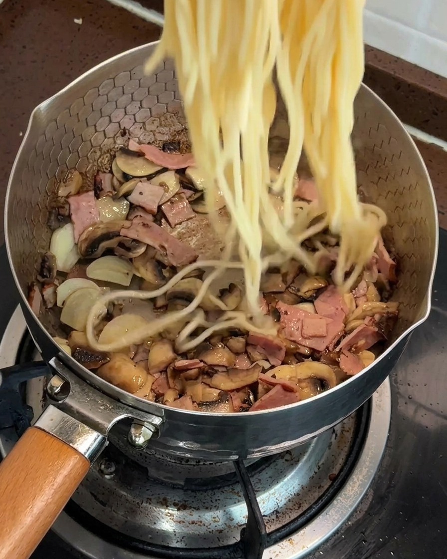 볶아진 버섯과 베이컨이 있는 팬에 삶은 스파게티 면을 넣는 모습.
