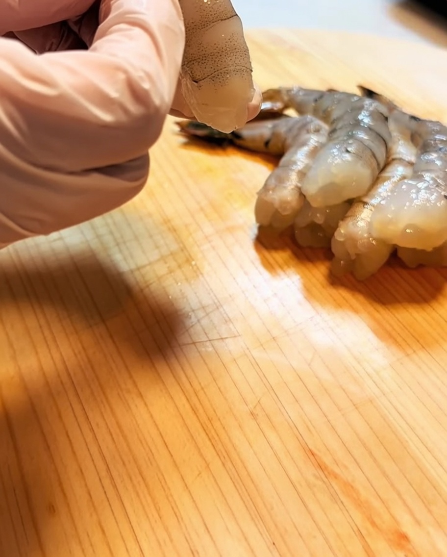 나무 도마 위에서 내장을 제거하고 손질된 새우를 핑크색 장갑을 낀 손으로 잡고 있는 모습.