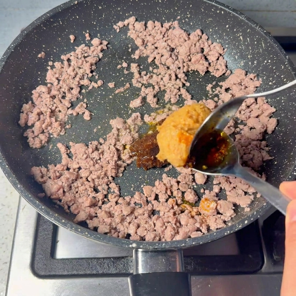 볶은 다진 소고기에 커다란 숟가락으로 걸쭉한 갈색 페이스트를 넣고 섞는 모습.
