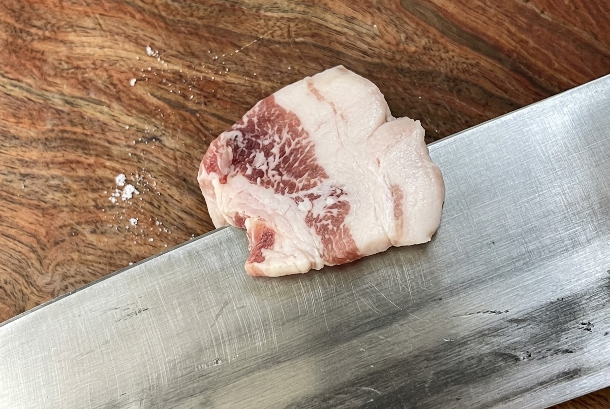 나무 도마 위에서 날카로운 칼로 생삼겹살을 얇고 균일하게 써는 모습.
