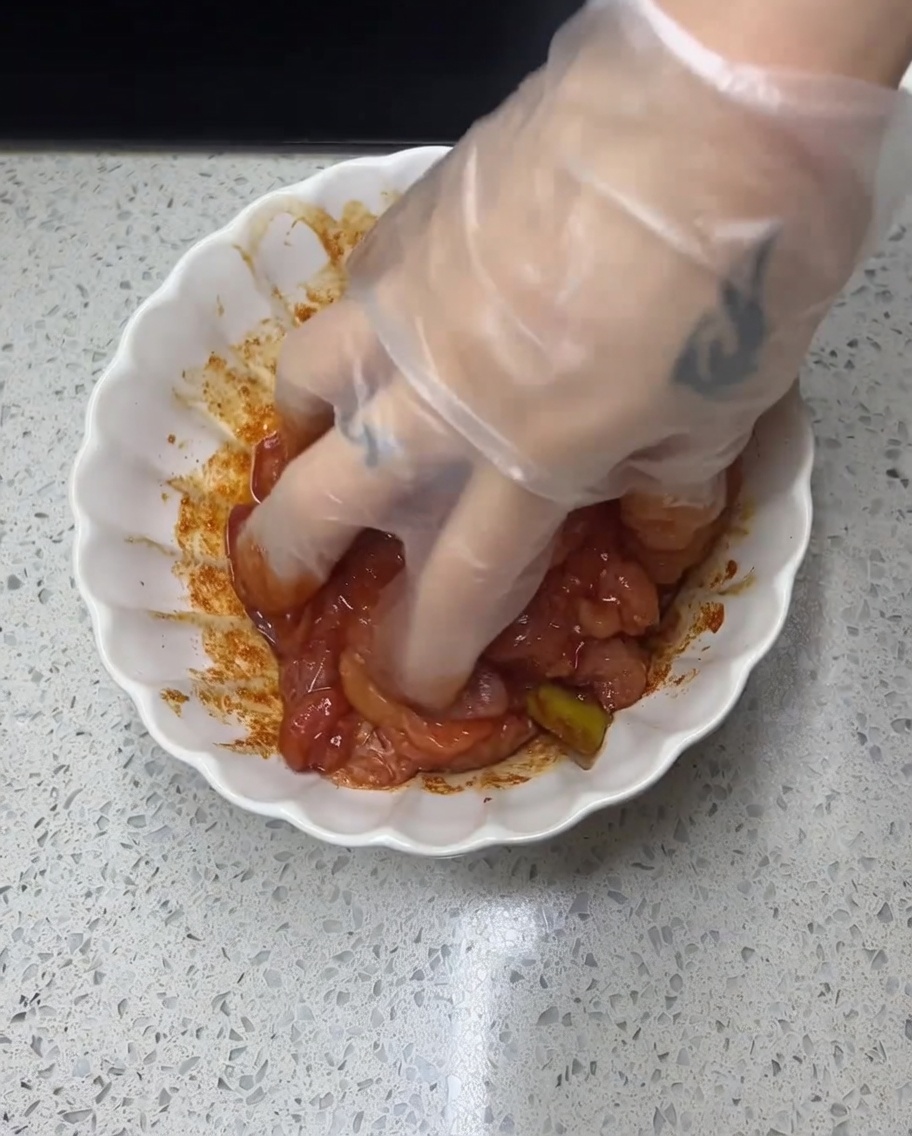 장갑을 낀 손으로 볼에 담긴 닭고기에 빨간 양념을 골고루 버무리는 모습.