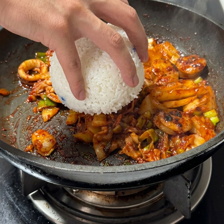 볶아진 김치와 고기가 있는 팬에 흰 쌀밥 한 공기를 넣는 모습.