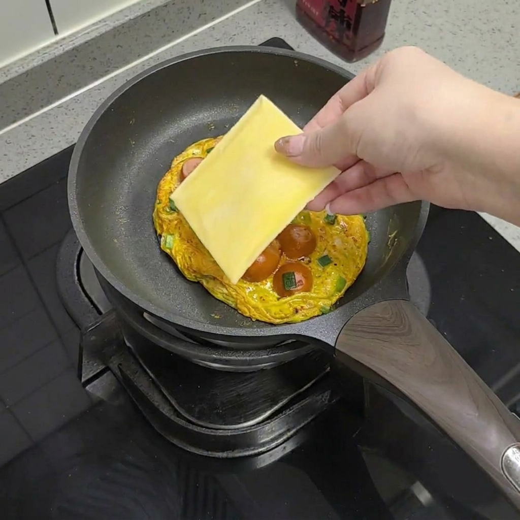 익은 계란과 소시지 위에 치즈 한 장을 올리는 모습.