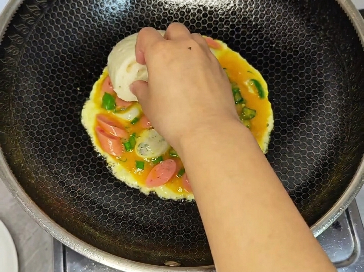 팬에서 익고 있는 젖은 계란과 소시지 혼합물 위에 흰 찐빵 반쪽을 손으로 누르는 모습.