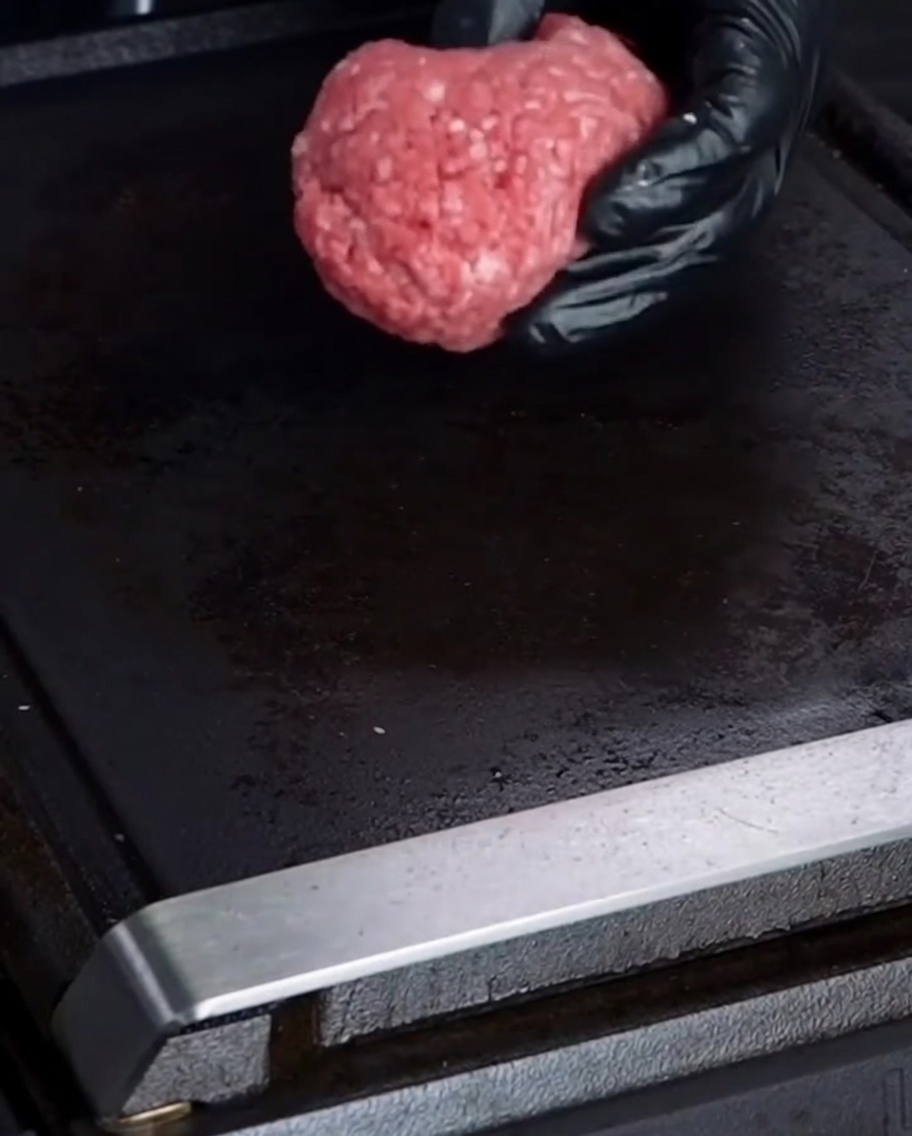 장갑 낀 손으로 소고기 반죽을 뜨거운 그릴 위에 올리는 모습.