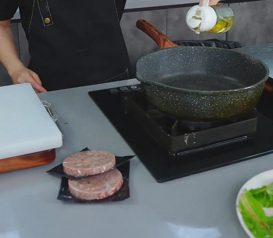 가스레인지 위에 놓인 어두운 색의 코팅 프라이팬에 소량의 식용유를 붓고 있는 모습.