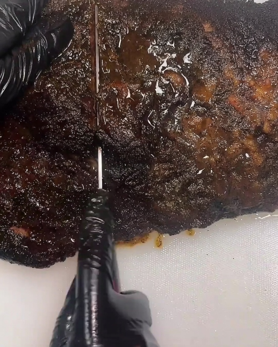 검은 장갑을 낀 사람이 긴 슬라이싱 칼로 훈연된 브리스킷을 써는 모습.