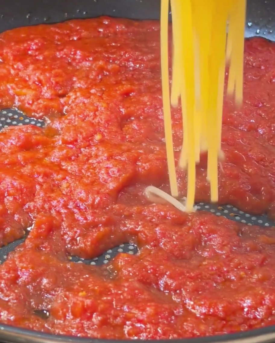 황금빛으로 잘 삶아진 스파게티 면을 들어 올려 진한 붉은색 토마토 소스가 담긴 팬으로 옮기는 모습.