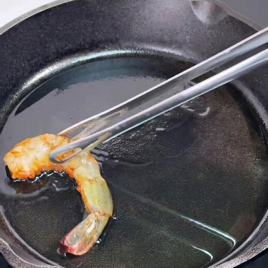 기름을 두른 뜨거운 무쇠 팬 위에서 주황빛으로 반쯤 익은 새우를 집게로 집어 올리는 모습.