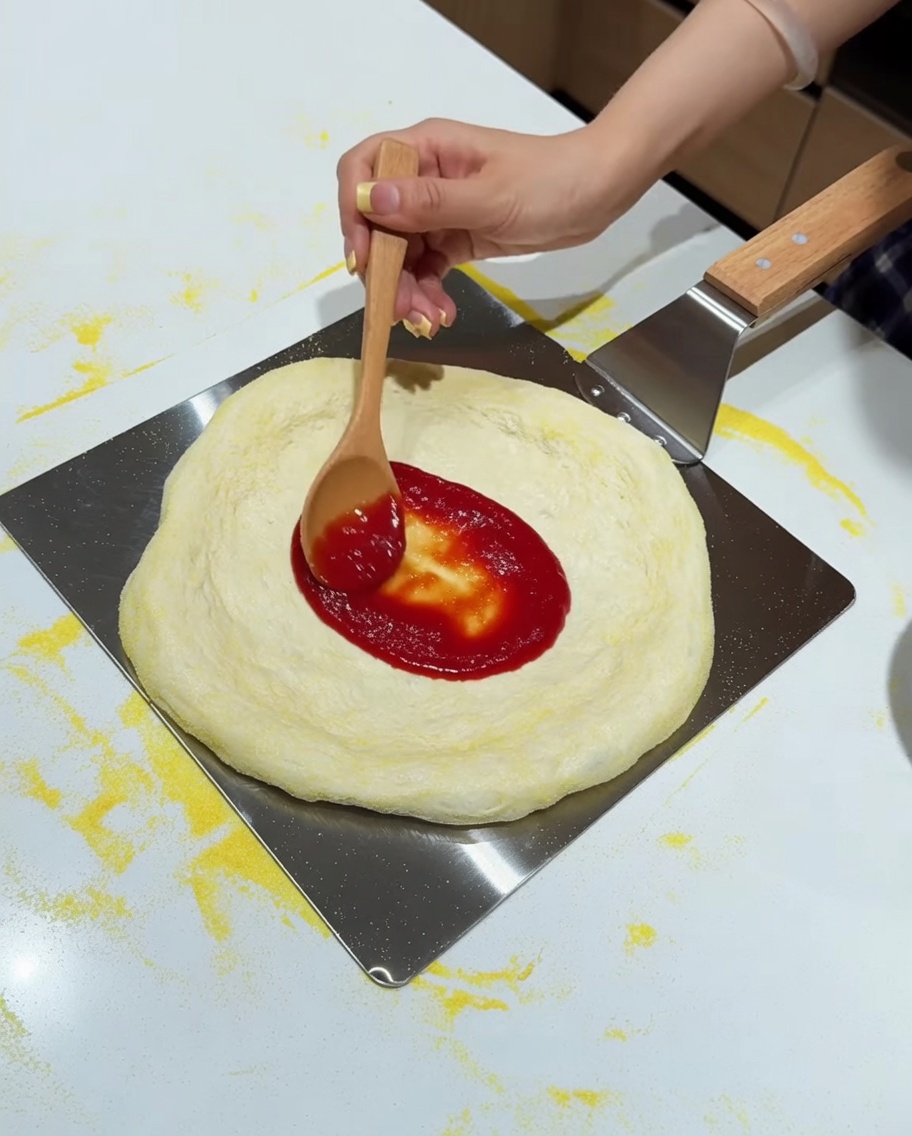 나무 숟가락으로 피자 도우에 토마토 소스를 펴 바르고 있습니다.