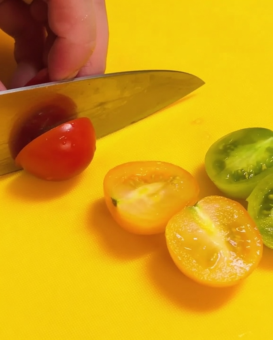 밝은 노란색 도마 위에서 빨간색과 초록색 방울토마토를 칼로 반으로 자르는 모습.