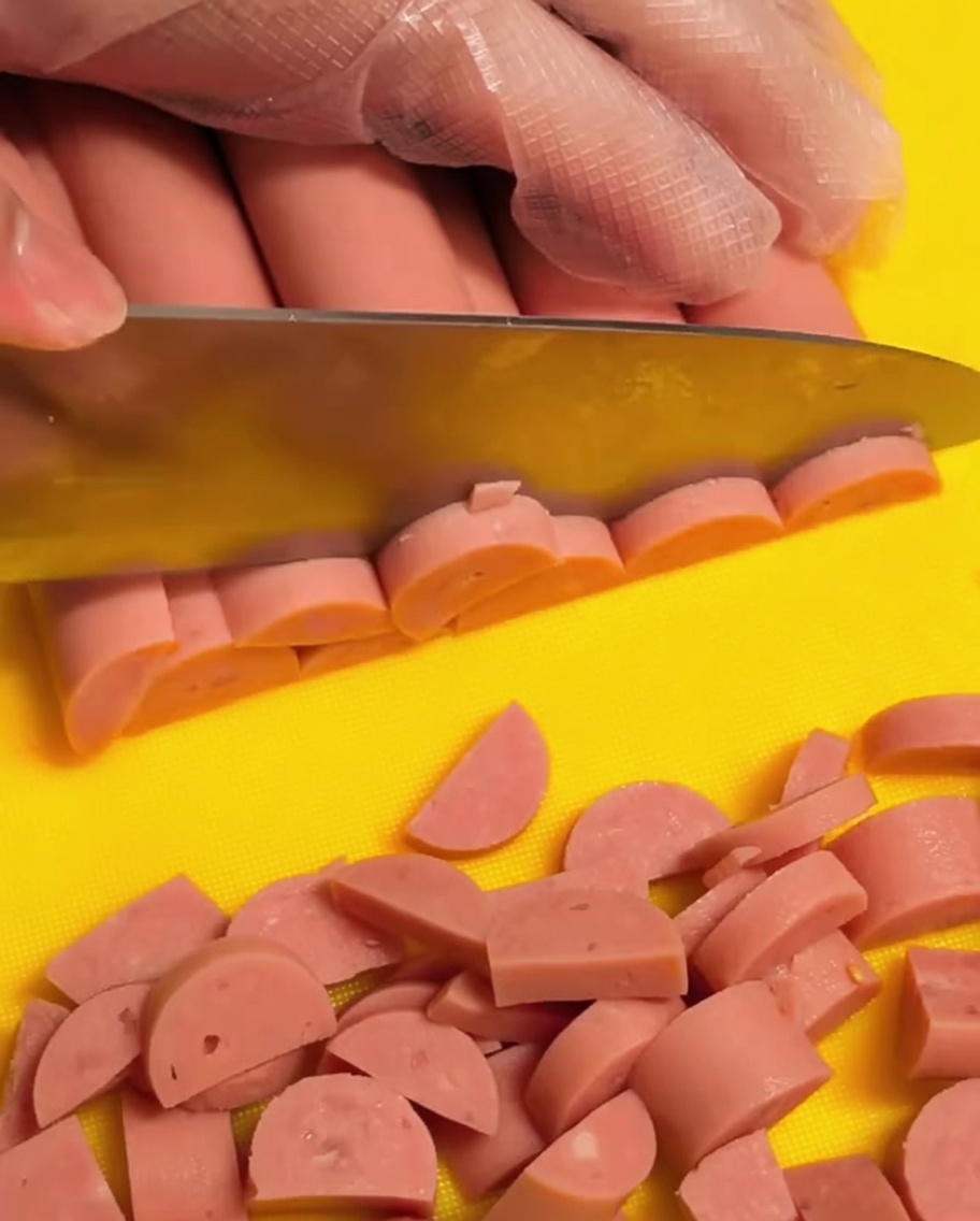 투명한 위생 장갑을 낀 손이 노란색 도마 위에서 분홍빛 햄 소시지를 반달 모양으로 작게 써는 모습.