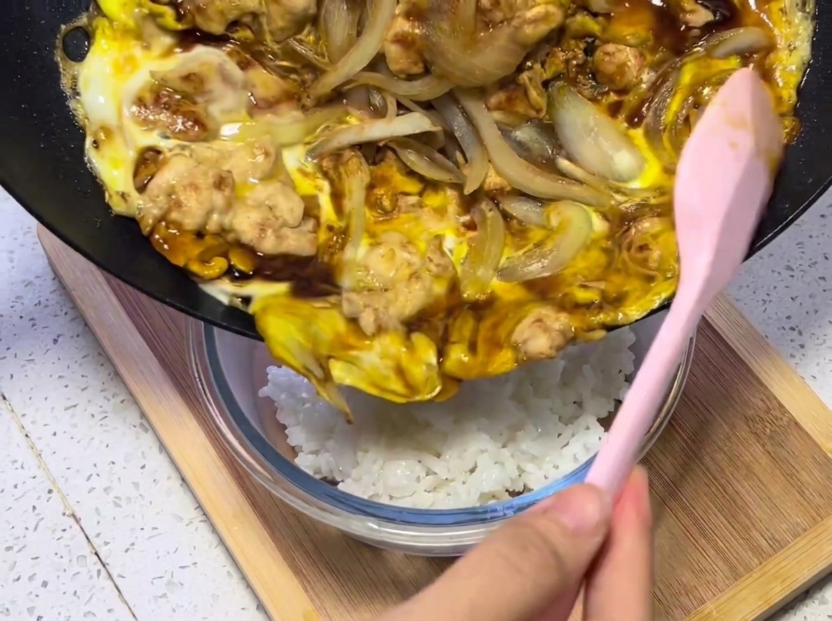 따뜻한 밥이 담긴 그릇 위에 팬에서 완성된 닭고기와 달걀 혼합물을 옮겨 담는 모습.