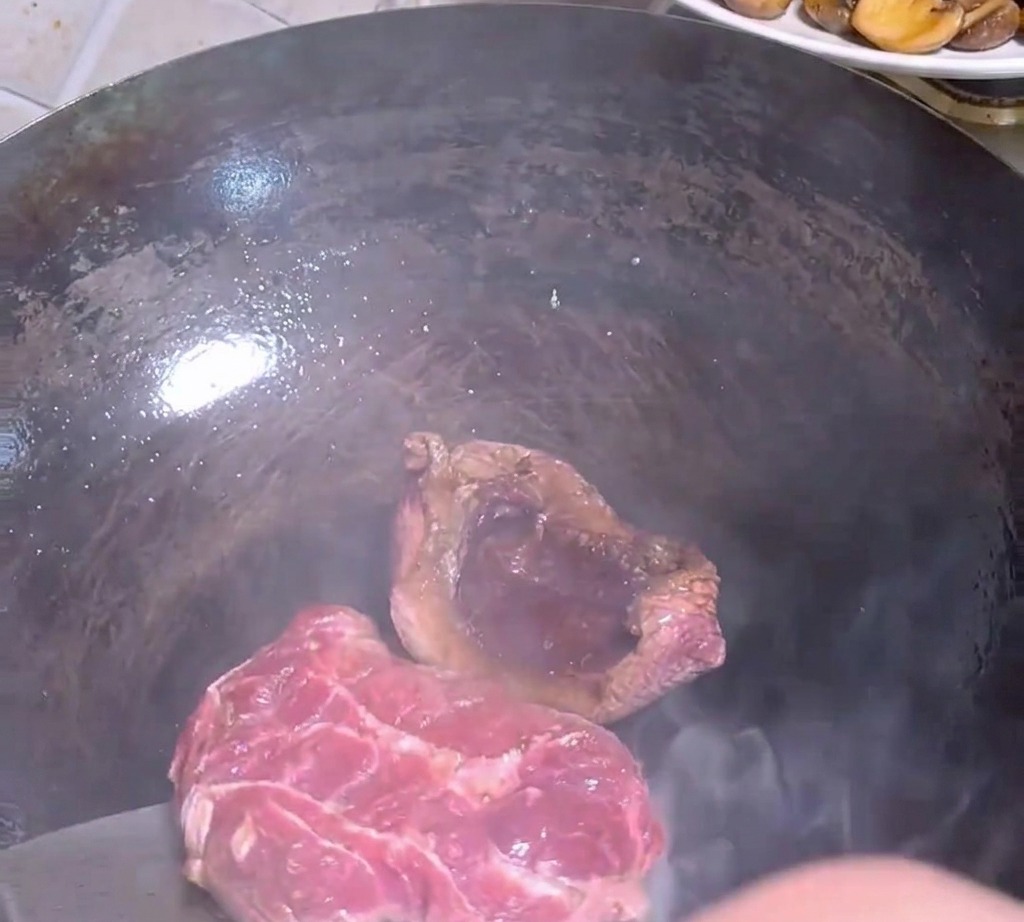 뜨겁고 어두운 색의 웍 안에서 쇠 주걱으로 겉이 구워진 스테이크를 뒤집는 모습.