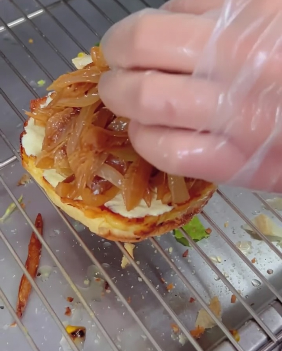 버거 번 위에 카라멜라이즈드 양파를 얹고, 그 위에 치즈가 녹은 소고기 패티를 층층이 쌓는 모습.
